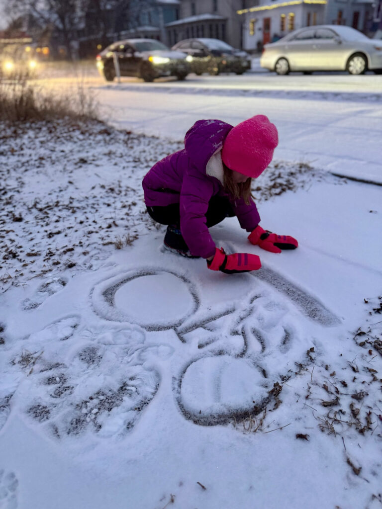 A young creative sees artistic potential in fresh snow; a child in pink gloves and a matching hat draws a person on a bike in the snow