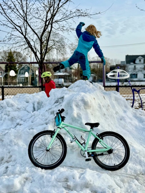 Two young kids leaping for joy over a snowbank with a bike in front of it