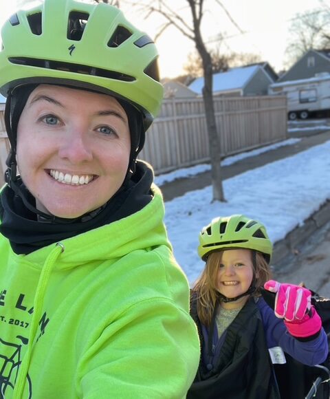 Laura Mitchell and one of her children take a selfie while commuting by bike in winter. Both riders wear bright neon helmets and big smiles of bike joy.